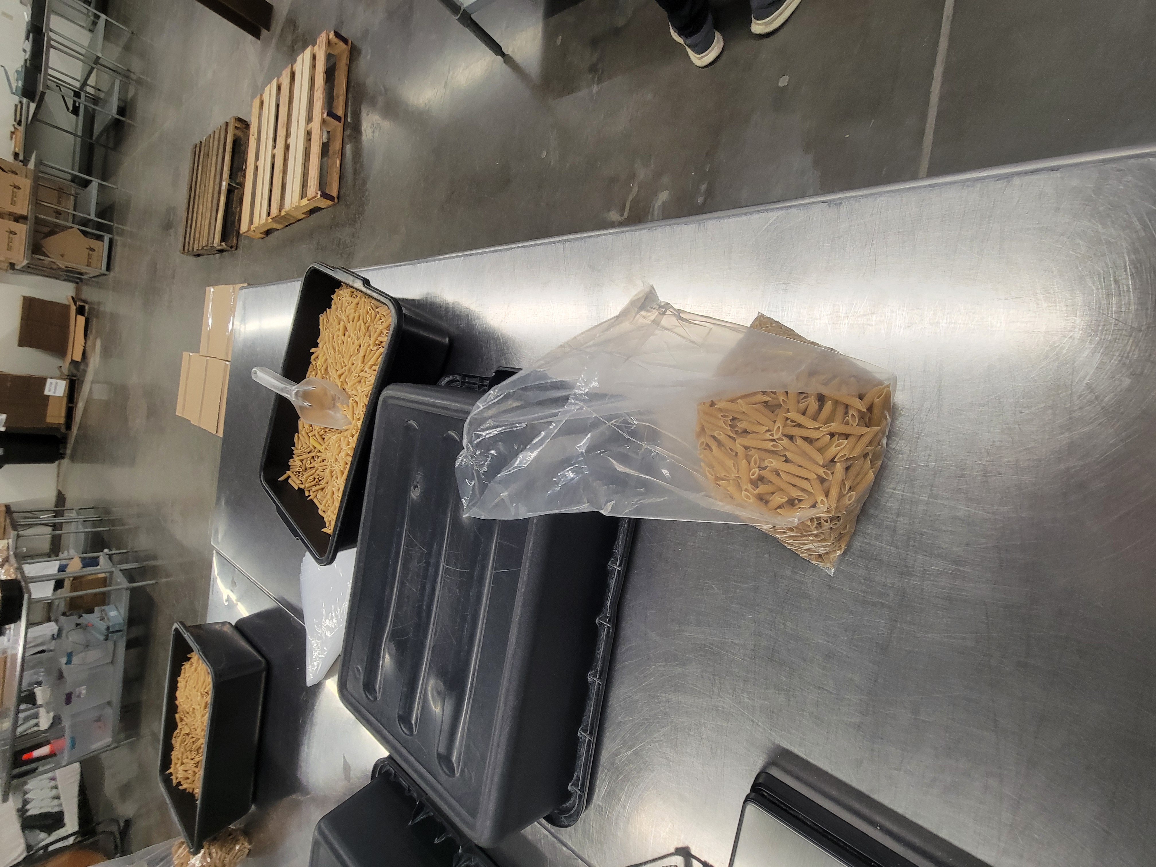 Bag of penne pasta with a tub flipped down and two other tubs full of penne pasta behind it.